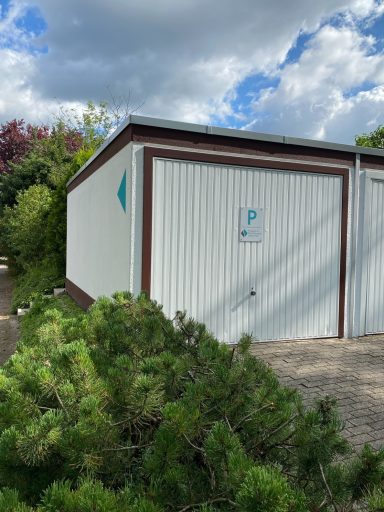 Weißes Gebäude mit Bäumen und blauem Logo an der Wand, unter einem wolkigen Himmel.